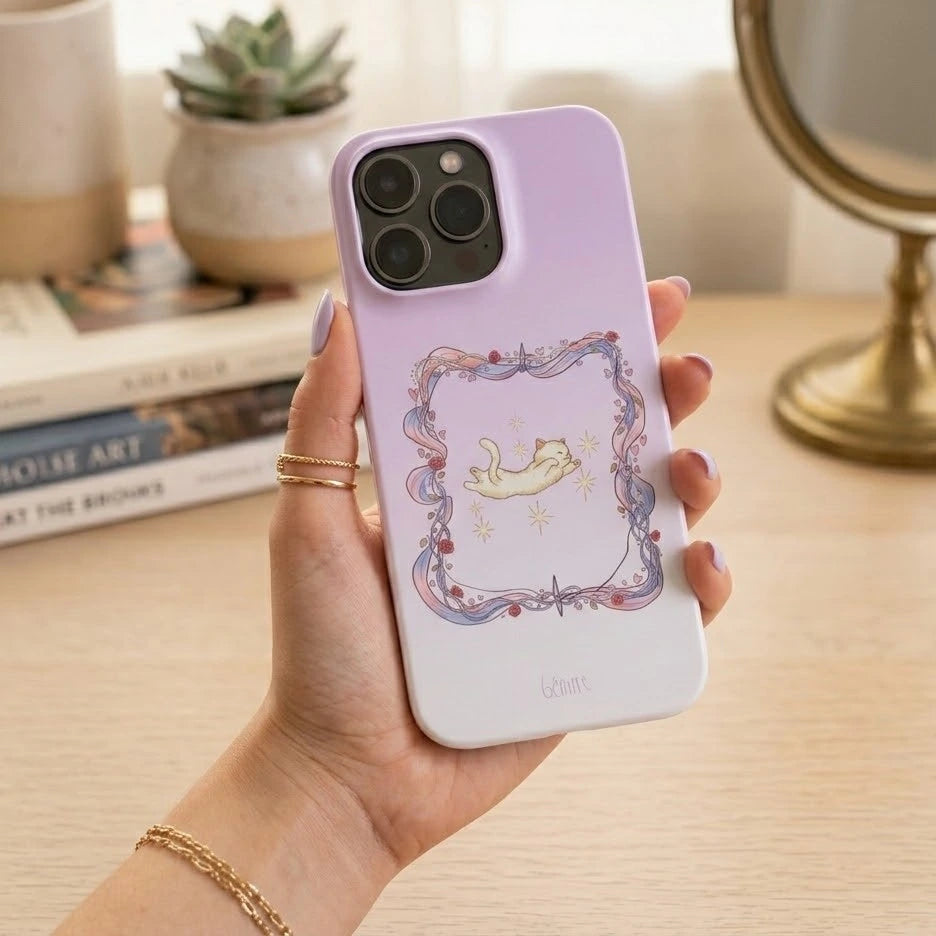 Hand holding a Bénirre phone case with a design of a cat in a romantic flower frame on a desk.